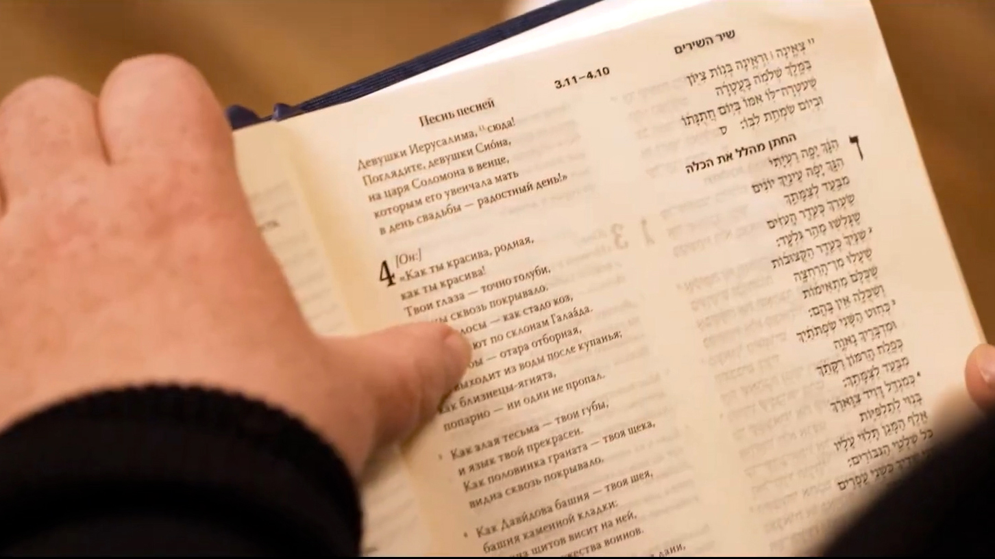 Hands holding an open Bible displaying bilingual text in Russian and Hebrew side-by-side.