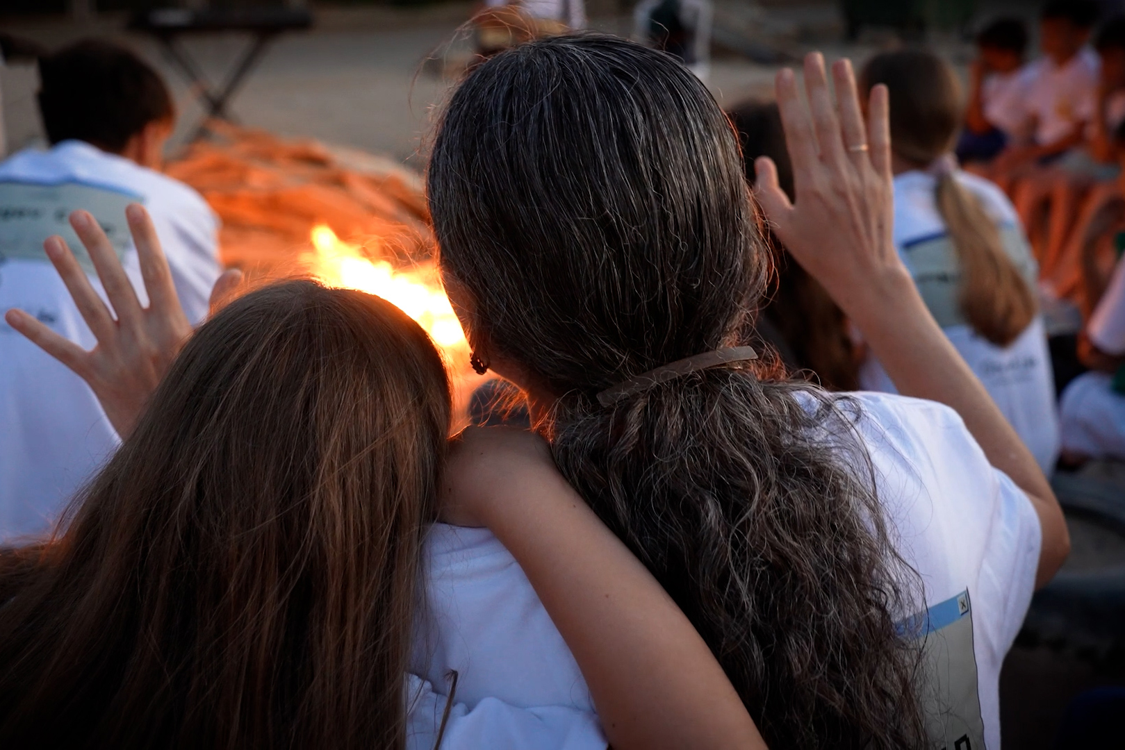 Back view of a woman and child with raised hands sitting around a warm outdoor campfire during a prayer service.