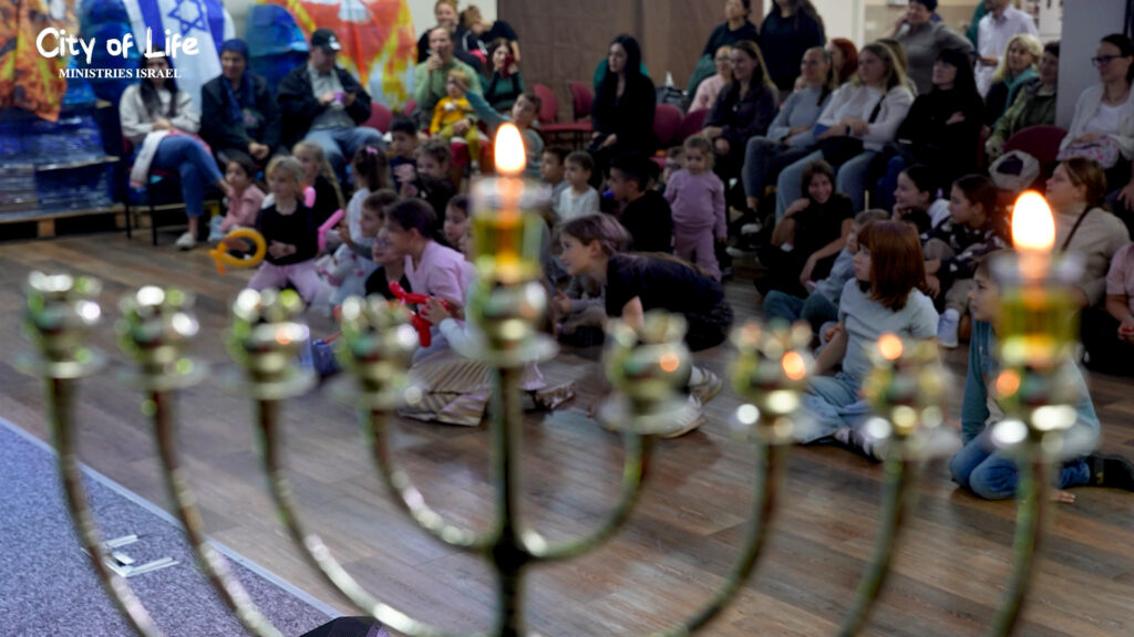 Menorah with lit candles in the foreground with families and children gathered in a hall behind it.